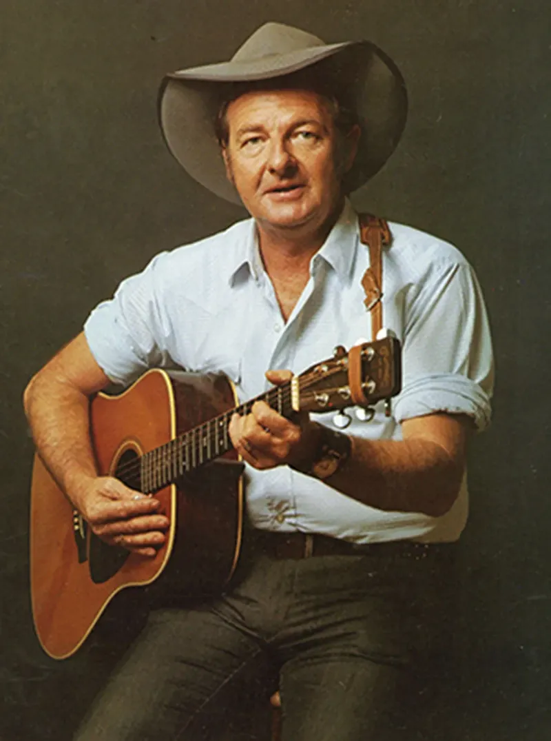 Slim Dusty in an Akubra hat and blue shirt, playing an acoustic guitar