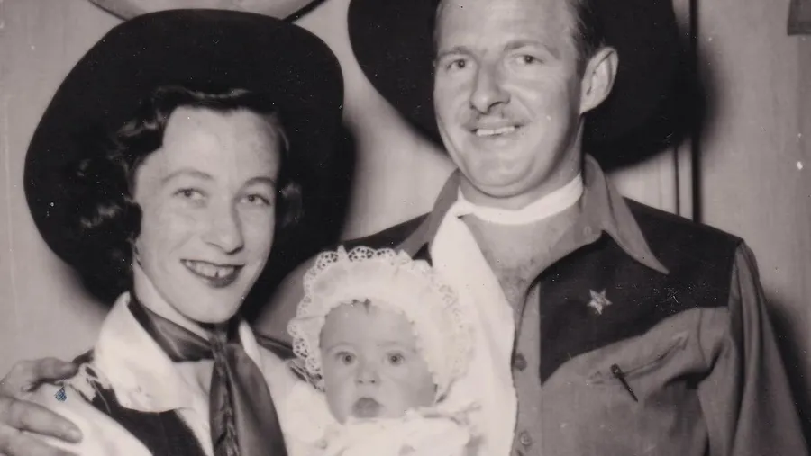 Joy McKean and Slim Dusty in western hats, smiling, holding a baby in a lace bonnet